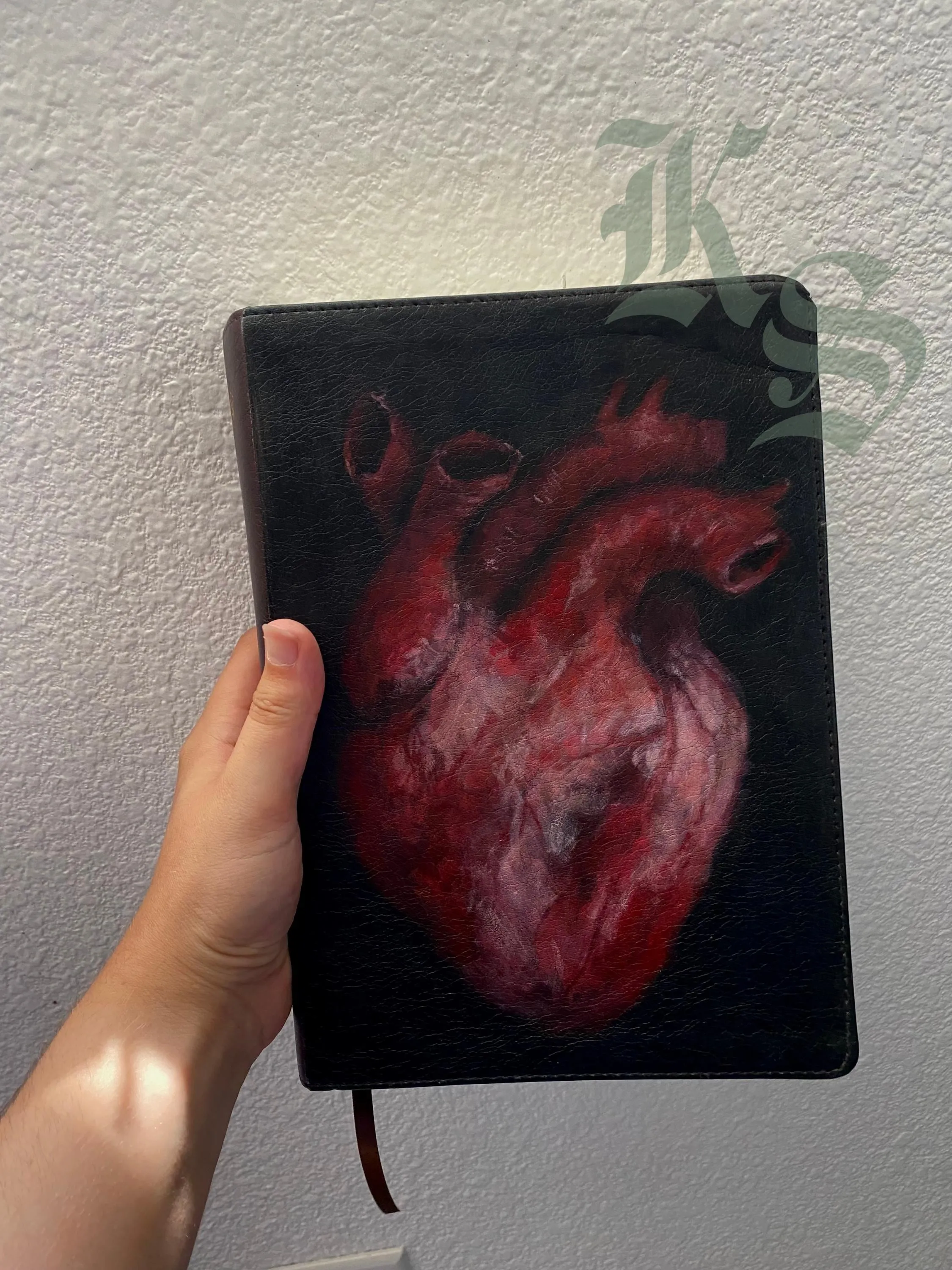 A hand holding a black leather ESV Bible with an anatomical heart hand-painted on the cover in deep reds.