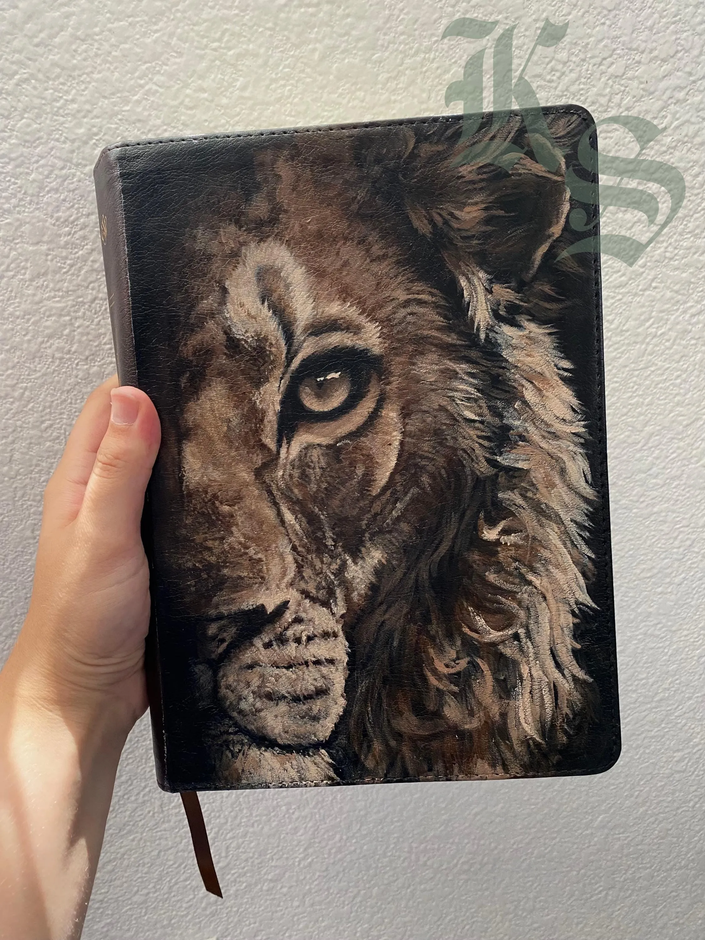 A hand holding a black leather ESV Bible with half a lion’s face hand-painted across the cover in warm browns.