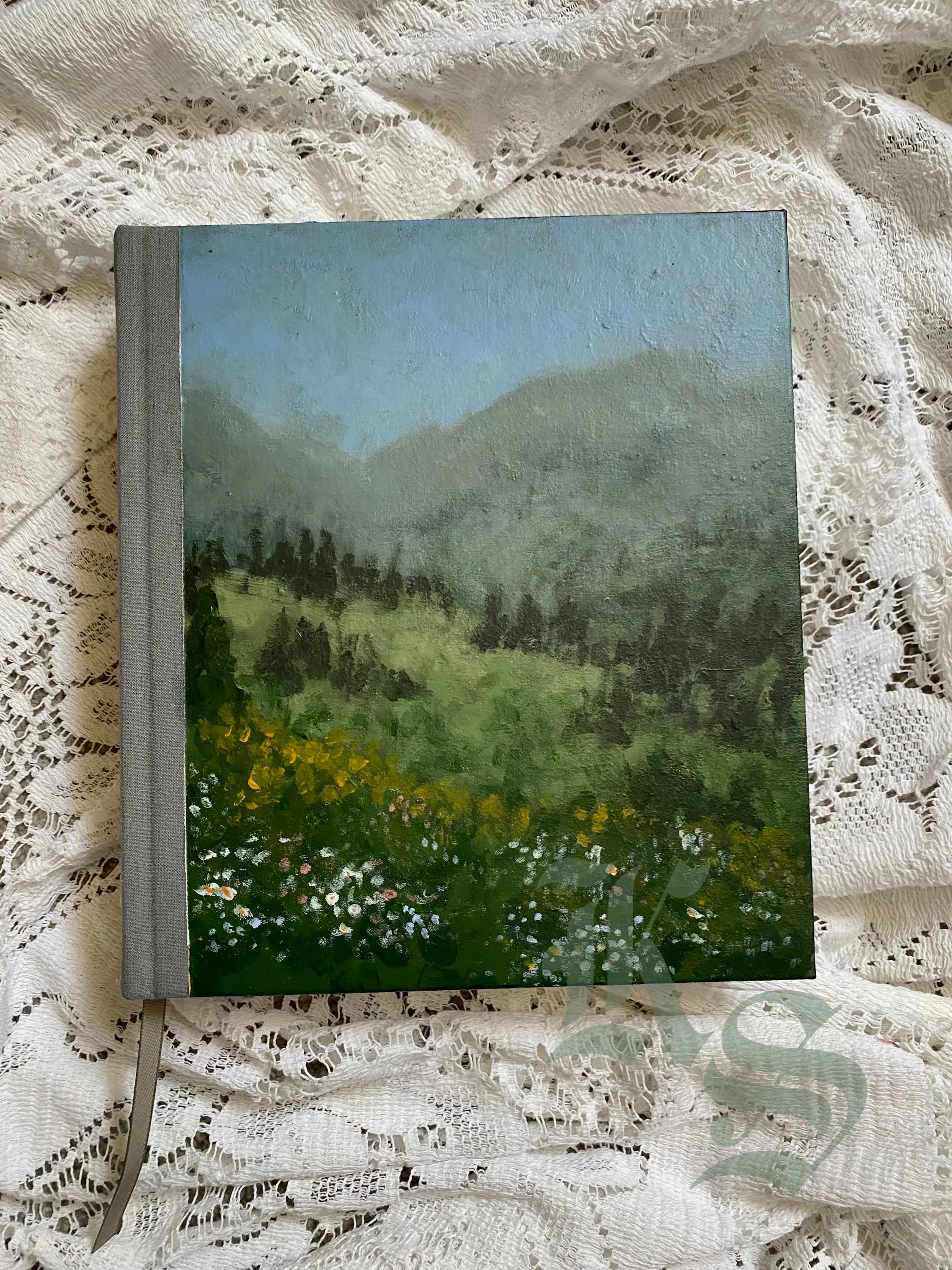 A grey hardcover ESV study Bible lying on lace, its cover painted with a green mountain meadow of wildflowers under a blue sky.