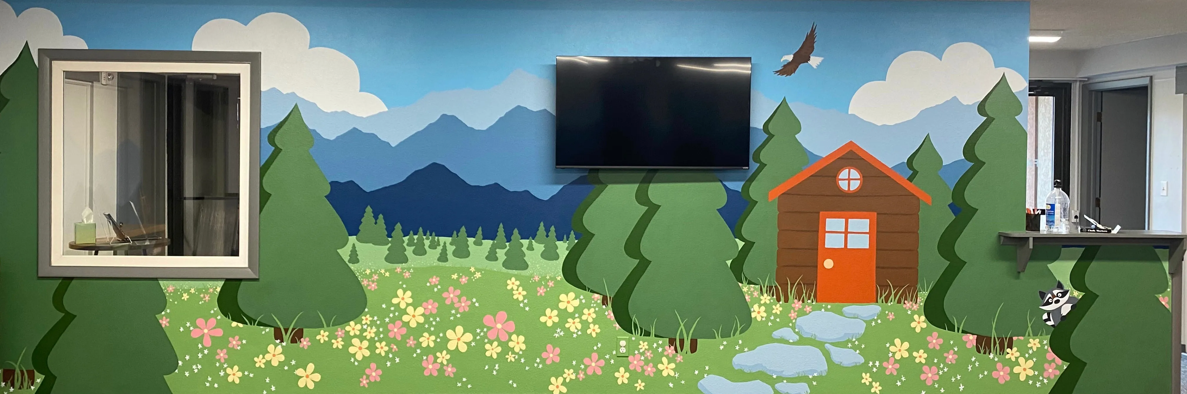 Painted wall mural in a children’s room: layered evergreen forest, a red cabin, a wildflower meadow and a stream, with blue mountains and a soaring bird beyond.
