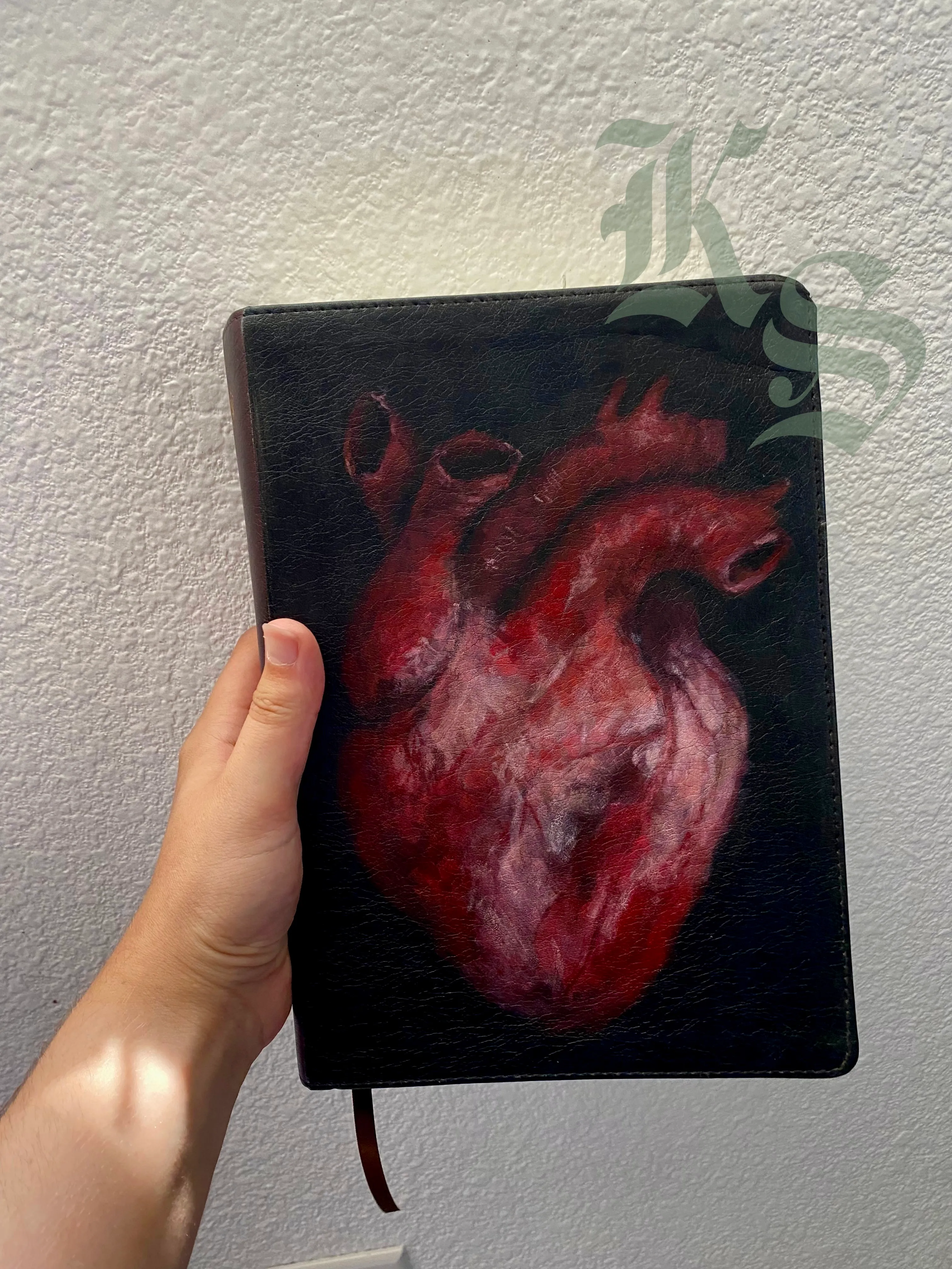 A hand holding a Bible with an anatomical heart hand-painted across the front cover in deep reds against black.