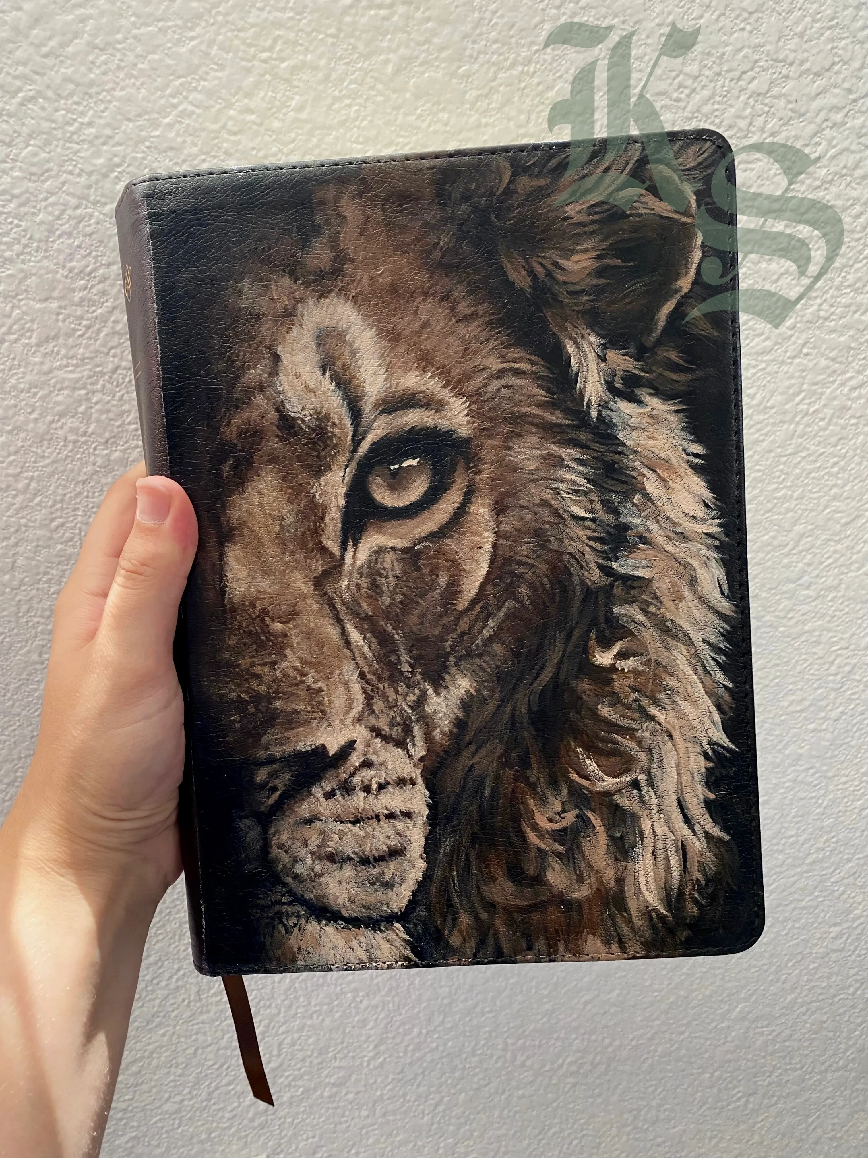 A hand holding a Bible with a lion’s face hand-painted across the front cover in warm browns.